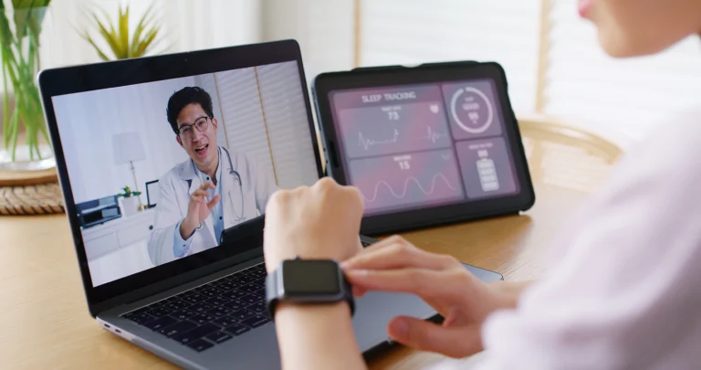 Patient receiving telehealth services from their doctor while wearing a smartwatch with fitness and health tracking abilities. There is also an external display next to their laptop showing analytics from their fitness tracker or health status.